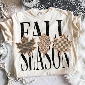 Może przedstawiać: Kremowy t-shirt z krótkim rękawem z czarnym napisem FALL SEASON. Na środku koszulki znajdują się trzy naszywki w kształcie liści, przedstawiające wzór moro, panterkę oraz beżowo-białą szachownicę.
