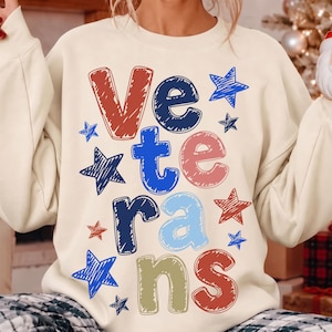 May include: A cream-colored crewneck sweatshirt featuring the word VETERANS printed in a multi-colored, scribbled-style font. The letters and surrounding star shapes are colored in shades of red, navy blue, light blue, and tan.