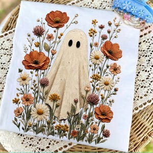 May include: A white t-shirt featuring a central beige ghost illustration surrounded by a detailed floral arrangement of orange, yellow, and brown poppies, daisies, and wildflowers with textured, raised-ink style graphic elements.
