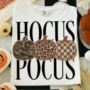 Op de afbeelding: Een wit t-shirt met de zwarte tekst HOCUS POCUS en drie pompoenen in het midden. De pompoenen hebben verschillende patronen: één met een groen en bruin camouflagepatroon, één met een bruine luipaardprint en één met een bruin dambordpatroon.