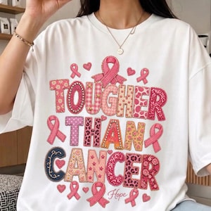 Peut inclure: Un t-shirt blanc à manches courtes avec le texte TOUGHER THAN CANCER en grandes lettres roses et rouges à motifs variés. Le design comprend divers symboles de rubans roses, des petits cœurs et le mot Hope en écriture cursive sous le texte.