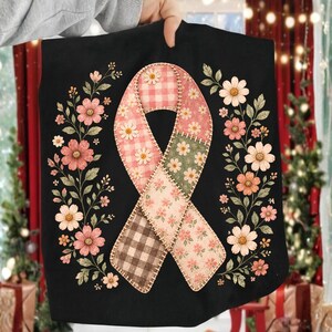 May include: A black fabric item featuring a central awareness ribbon graphic composed of various patchwork patterns including pink gingham, green floral, and brown plaid. The ribbon is flanked by two vertical floral vines with pink flowers.