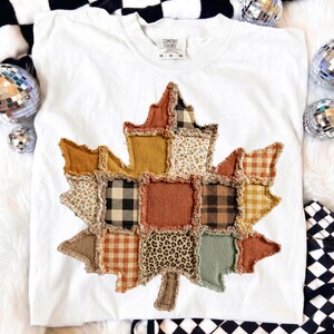 May include: A white short-sleeve t-shirt featuring a large maple leaf applique made from various fabric squares with frayed edges. The patches include patterns like plaid, gingham, floral, and leopard print in shades of orange, brown, and green.