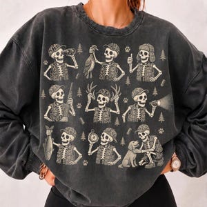 May include: A charcoal gray crewneck sweatshirt featuring a graphic print of nine skeletons dressed in hunting gear. The skeletons are depicted performing various outdoor activities including holding a rifle, a fish, a duck, a compass, and a dog.