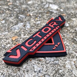 Puede incluir: Un emblema impreso en 3D negro y rojo con la palabra "VICTORY" y "MOTORCYCLES" en letras rojas y "USA" en letras blancas.