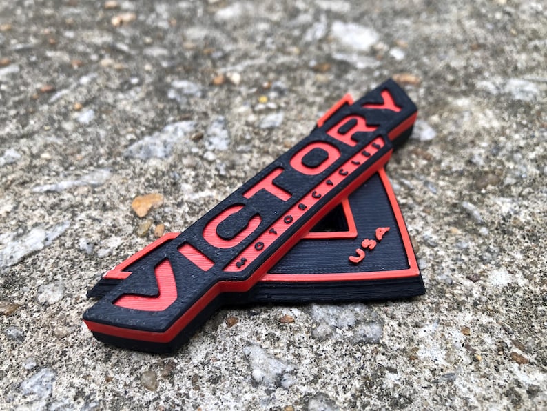 Puede incluir: Un emblema impreso en 3D negro y rojo con la palabra "VICTORY" y "MOTORCYCLES" en letras rojas y "USA" en letras blancas.