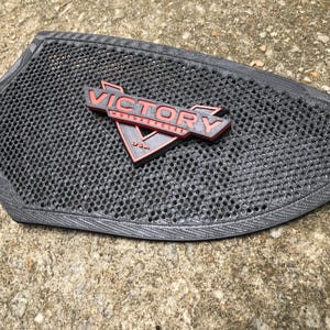 Puede incluir: Un asiento de motocicleta negro con un logotipo de Victory Motorcycles negro y rojo. El asiento tiene un patr&oacute;n de panal.