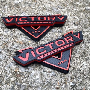 Puede incluir: Dos logotipos de Victory Motorcycles impresos en 3D en negro y rojo. Los logotipos tienen forma de tri&aacute;ngulo con la palabra "Victory" en letras rojas y "Motorcycles" en letras rojas m&aacute;s peque&ntilde;as debajo. La palabra "USA" tambi&eacute;n est&aacute; en letras rojas en la parte inferior del tri&aacute;ngulo.