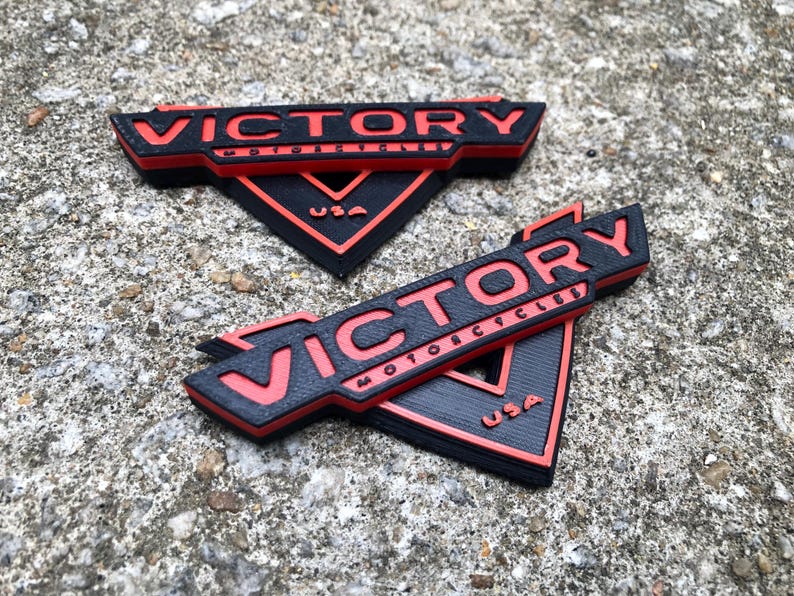 Puede incluir: Dos logotipos de Victory Motorcycles impresos en 3D en negro y rojo. Los logotipos tienen forma de tri&aacute;ngulo con la palabra "Victory" en letras rojas y "Motorcycles" en letras rojas m&aacute;s peque&ntilde;as debajo. La palabra "USA" tambi&eacute;n est&aacute; en letras rojas en la parte inferior del tri&aacute;ngulo.