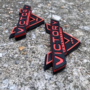 Puede incluir: Dos logotipos de Victory Motorcycles impresos en 3D en negro y rojo. Los logotipos tienen forma triangular con la palabra "Victory" en rojo y "Motorcycles" en negro. Los logotipos est&aacute;n sobre una superficie de hormig&oacute;n gris.