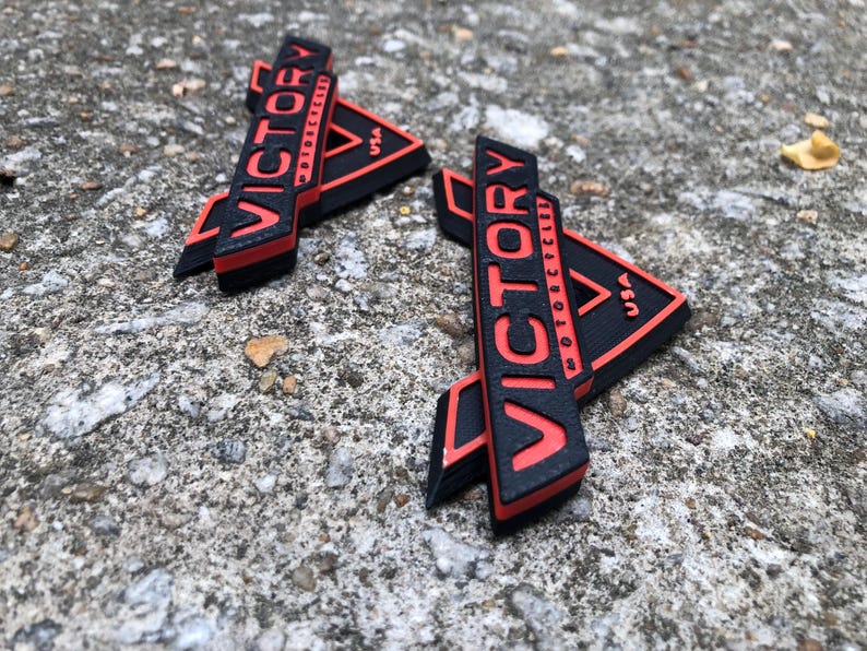 Puede incluir: Dos logotipos de Victory Motorcycles impresos en 3D en negro y rojo. Los logotipos tienen forma triangular con la palabra "Victory" en rojo y "Motorcycles" en negro. Los logotipos est&aacute;n sobre una superficie de hormig&oacute;n gris.