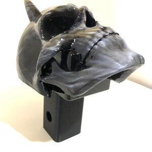May include: A black and white skull hitch cover with a single horn. The skull is detailed with a black and white marbled paint effect. The hitch cover is attached to a black metal hitch receiver.