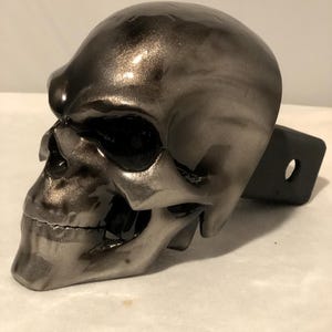 May include: A metallic silver skull-shaped trailer hitch cover. The skull has detailed features, including eye sockets, a nose cavity, and a jawline. The hitch cover is designed to fit a standard trailer hitch receiver.