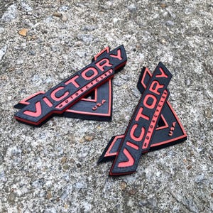 Puede incluir: Dos emblemas triangulares negros y rojos con la palabra "VICTORY" en letras rojas y las palabras "MOTORCYCLES" y "USA" en letras blancas m&aacute;s peque&ntilde;as.