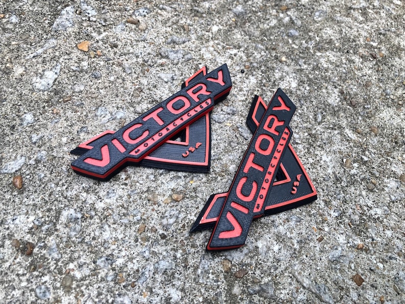 Puede incluir: Dos emblemas triangulares negros y rojos con la palabra "VICTORY" en letras rojas y las palabras "MOTORCYCLES" y "USA" en letras blancas m&aacute;s peque&ntilde;as.