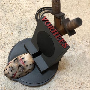 May include: A black phone stand featuring a weathered hockey mask with red markings, two upright knife handles, and the red text VORHEES embossed vertically on the side. The stand includes a circular base and a black power cord extending from the back.