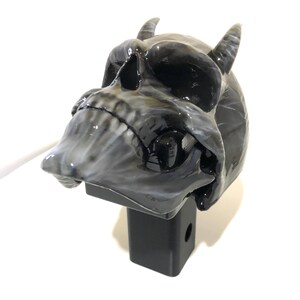 May include: A black and white skull with horns attached to a black hitch receiver. The skull has a glossy finish.