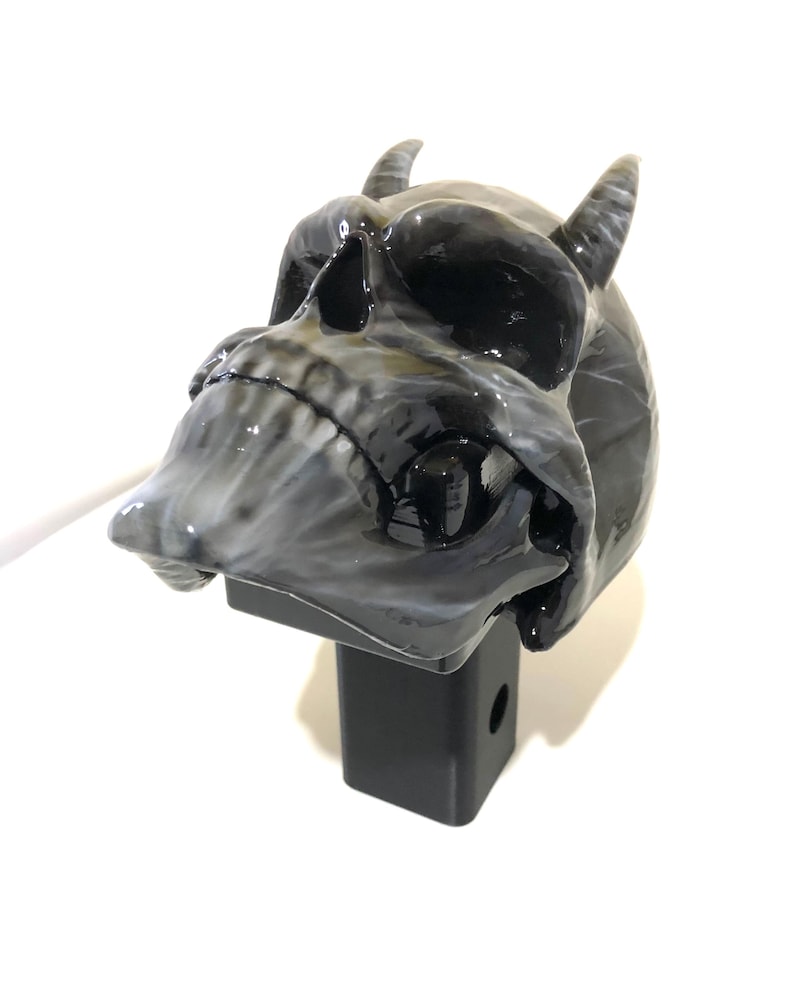 May include: A black and white skull with horns attached to a black hitch receiver. The skull has a glossy finish.