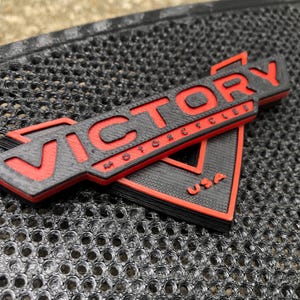 Puede incluir: Un logotipo de Victory Motorcycles impreso en 3D en negro y rojo con el texto "VICTORY MOTORCYCLES USA".