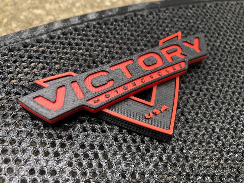 Puede incluir: Un logotipo de Victory Motorcycles impreso en 3D en negro y rojo con el texto "VICTORY MOTORCYCLES USA".