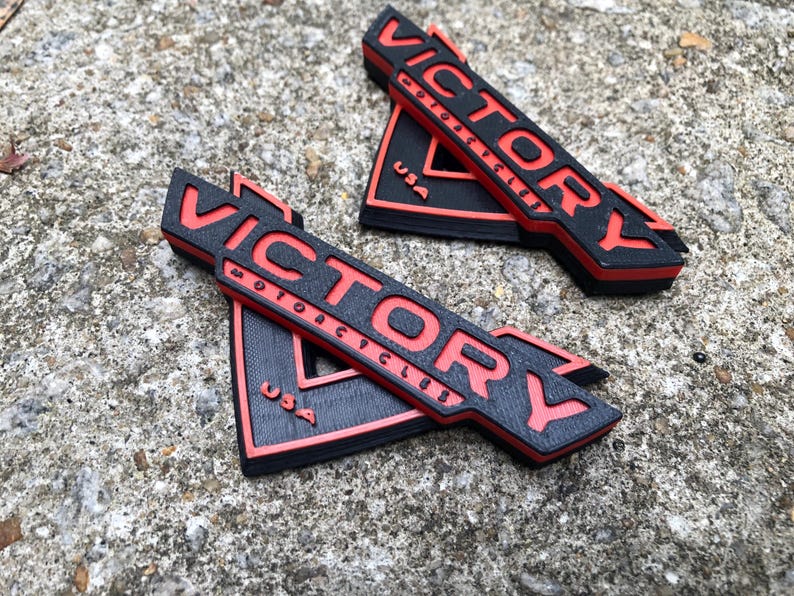 Puede incluir: Dos emblemas impresos en 3D en negro y rojo con el texto "VICTORY MOTORCYCLES USA" en ellos.