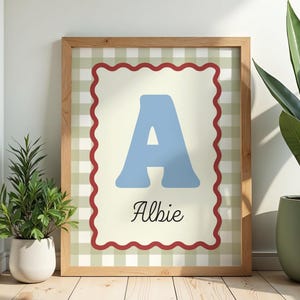 May include: A framed print with a blue letter "A" and the name "Albie" in script. The print has a red scalloped border and a green and white checked background. The frame is made of light-coloured wood, suitable for a child's room.