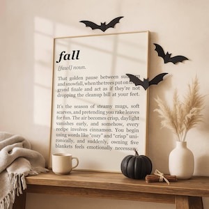 Könnte beinhalten: Ein gerahmter Kunstdruck mit der Definition des Wortes fall, schwarzer Text auf cremefarbenem Hintergrund. Der Druck wird mit drei schwarzen Papierfledermäusen, einer kleinen Keramiktasse, einem schwarzen Kürbis und einer Vase mit Pampasgras gezeigt.