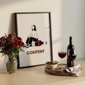 May include: A framed wall art print featuring a black and white line drawing of a person sitting on a large red wine bottle. The text WINE is printed on the bottle label, and the word CONTENT is printed in dark red below the illustration.