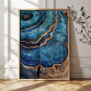 May include: A framed print featuring a close-up of a tree stump with concentric rings. The artwork showcases a striking blend of deep blue and gold hues, with intricate details of the wood grain. The frame is a light brown color.