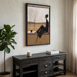 Puede incluir: Impresión artística enmarcada que representa a una persona con un vestido negro leyendo junto a una ventana. La obra de arte presenta una paleta de colores apagados y un marco negro. Una taza de café está en el alféizar. El arte está sobre un mueble de madera oscura.