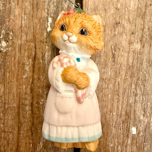 May include: A ceramic cat figurine with orange fur, blue eyes, and a pink bow. The cat wears a pink dress with a blue trim and holds a candy cane. A wire loop is attached for hanging. The figurine is set against a wooden background.