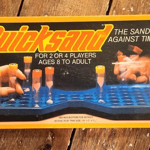 Könnte beinhalten: Eine gelbe Schachtel mit dem Spiel "Quicksand: The Sand Race Against Time". Das Spiel ist für 2 bis 4 Spieler, ab 8 Jahren bis Erwachsene. Die Schachtel zeigt ein blaues Spielbrett mit Sanduhren und eine Hand, die nach einer Sanduhr greift.