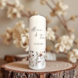 Könnte beinhalten: Eine weiße Stumpenkerze mit den Namen "Alina & Tim" und dem Datum "28. Mai 2025" in schwarzer Schrift. Der Fuß der Kerze ist mit einem floralen Muster in Rosa, Lila und Gelb verziert. Die Kerze steht auf einer Holzscheibe.