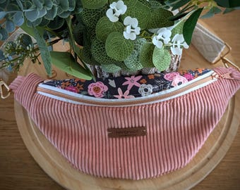 May include: A pink corduroy fanny pack with a floral patterned zipper pocket. The bag has a gold zipper and hardware. The word "HANDMADE" is on a small brown tag on the front of the bag. The bag is on a wooden surface.