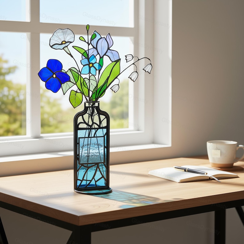 May include: A stained glass vase with a bouquet of colorful flowers. The flowers include blue, white, and green blooms. The vase is black with blue glass and has the text "Jackson's Garden". The vase is on a wooden table.