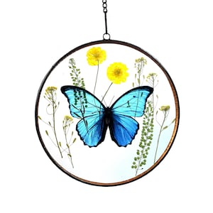 May include: A blue morpho butterfly with yellow flowers and green foliage encased in a round glass ornament with a black metal frame and chain.