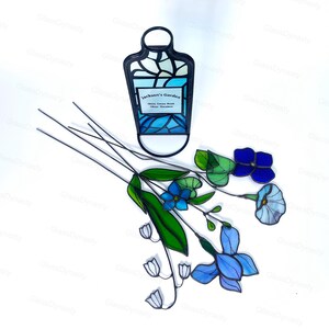 May include: Stained glass art piece featuring a decorative bottle shape with the text "Jackson's Garden". The bottle is in shades of blue and white. Also, a collection of stained glass flowers in blue, green, and white.