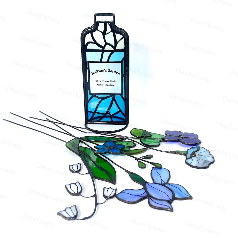 May include: A stained glass bottle-shaped piece with the text "Jackson's Garden" and names. It features blue and white glass, with a black metal frame. Several stained glass flowers in shades of blue, purple, and green are also present.