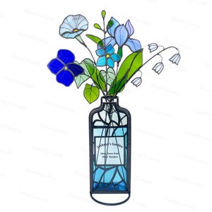 May include: Stained glass floral arrangement in a vase. The flowers are blue, green, and white. The vase is a rectangular shape with a black frame and a blue interior. The vase has the text "Jackson's Garden" and names.