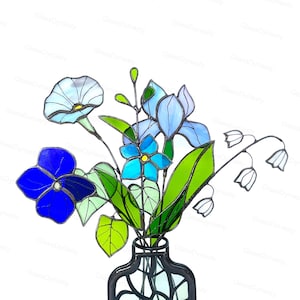 May include: A stained glass bouquet featuring blue and white flowers, green leaves, and white lily of the valley blooms. The flowers are arranged in a black vase. The image is set against a white background.