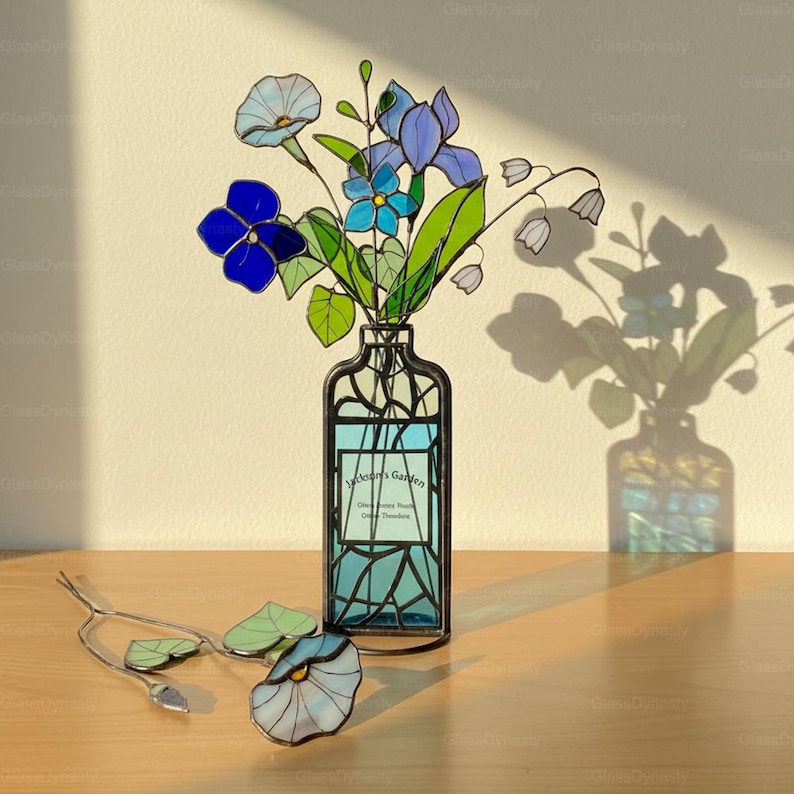 May include: A stained glass vase with blue and purple flowers, green leaves, and a stem. The vase is rectangular with a black frame and a label that reads "Jackson's Garden". The vase is on a wooden surface.