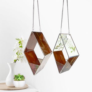 May include: Two diamond-shaped stained glass hanging planters with silver chains. One planter has a brown and iridescent glass design, the other has a clear glass design with dried white flowers.