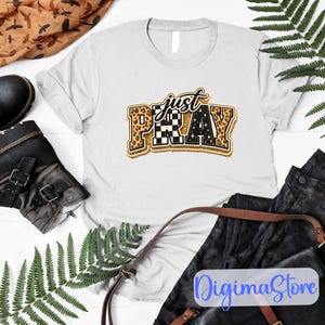 Könnte beinhalten: Weißes T-Shirt mit dem Schriftzug "just PRAY" in einem dekorativen Design. Das Wort "just" ist in goldener Schrift geschrieben, und "PRAY" ist in einem Blockbuchstabenstil mit Leoparden-, Schachbrett- und Sternmustern gehalten. Das Shirt ist auf einer weißen Oberfläche gefaltet.