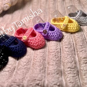 May include: A collection of six crocheted baby shoes in various colors, including black, navy, pink, purple, yellow, and gray. Each shoe features a strap with a button closure. The text "CrochetbyTamisha" is visible in the image.