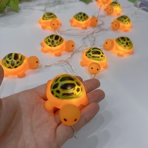 May include: A string of orange turtle-shaped LED lights. Each turtle has a patterned green and black shell and a smiling face. The lights are strung on a thin, clear wire, ideal for decorative use.