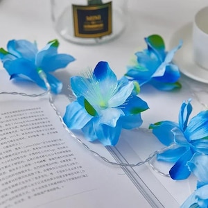 May include: A string of blue artificial flower lights. Each flower has multiple petals in shades of blue and green, with a cluster of white fiber optic strands in the centre. The lights are on a thin silver wire.