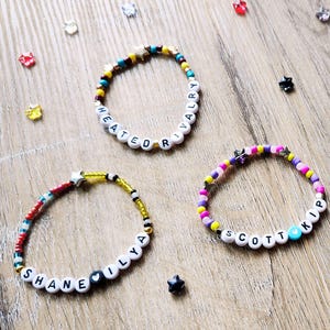 Może przedstawiać: Trzy bransoletki z koralików z białymi koralikami literowymi, które wymawiają imiona. Bransoletki są wielokolorowe z akcentami żółtego, niebieskiego i różowego. Rozrzucone są gwiazdki i inne ozdobne koraliki. Bransoletki leżą na drewnianej powierzchni.