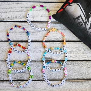 Peut inclure: Cinq bracelets en perles colorées avec des perles de lettres blanches qui épellent différents mots. Les bracelets sont sur une surface en bois. Les mots sur les bracelets sont "Hammertime", "Shadger", "Honeybee", "Incident", et "Alexbonbo".