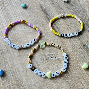 May include: Three colorful beaded bracelets with different words spelled out on them. The bracelets are on a wooden surface with small heart-shaped beads scattered around them. The words on the bracelets are "Tough Love", "Risk", and "I'm Sorry".