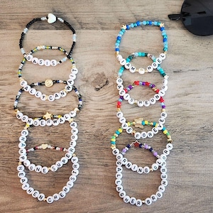 Puede incluir: Una colección de pulseras de cuentas coloridas con diferentes palabras escritas en ellas. Las pulseras están apiladas unas sobre otras en una superficie de madera. Las palabras en las pulseras incluyen "Oasis", "Sally", "Can Wait", "Wonderwall", "Champagne", "Supernova", "Heathen", "Chemistry", "I Am", "Noel", "Oasis Reunion", "Live Forever", "Morning", "Gallagher", "Brother", and "Glory".